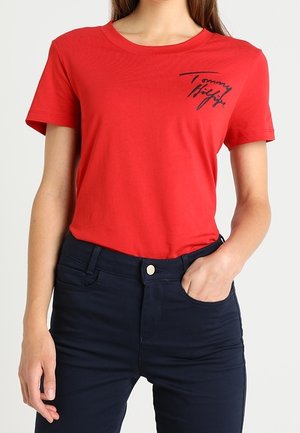 Kobieta ubrana w czerwoną koszulkę Tommy Hilfiger i ciemnoniebieskie spodnie z wysokim stanem, z jedną dłonią w kieszeni, na jednolitym tle.