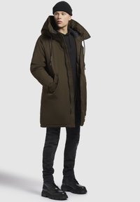 Młody mężczyzna w czarnym beanie, długim oliwkowym parku, czarnej bluzie z kapturem, ciemnych dżinsach i czarnych masywnych butach, stojący na jednolitym tle.