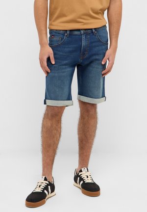 Homme portant un short en jean bleu retroussé, une chemise beige et des baskets noires à lacets blancs, debout devant un fond clair uni.