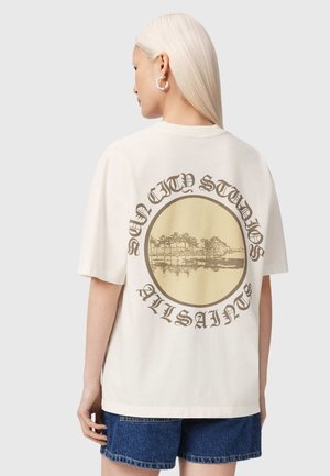 Kvinde med langt blondt hår iført en cremefarvet t-shirt med en rund palme træ-grafik og teksten "Sun City Studios AllSaints" på ryggen.