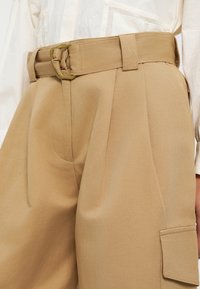Pantalon beige sur mesure avec ceinture nouée, doté de poches latérales et d'une coupe structurée, fabriqué dans un tissu lisse avec une texture subtile.