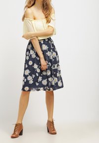 Żółty top odsłaniający ramiona zestawiony z granatową spódnicą w białe wzory stokrotek. Brązowe buty na obcasie ze sznurówkami. Gładka tekstura tkaniny.