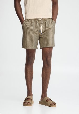 Mężczyzna w beżowych i czarnych kraciastych szortach ze sznurkiem oraz beżowych zamszowych sandałach, stoi z rękami w kieszeniach na białym tle.