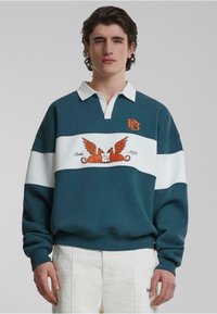 Pulover teal cu dungi albe și albastre deschise, având un logo brodat portocaliu și o grafică decorativă cu două creaturi cu aripi.