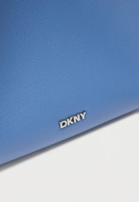Modrá kožená kabelka s textúrou, na ktorej je strieborné logo DKNY. Dizajn zahŕňa hladký povrch a materiál vysokej kvality.