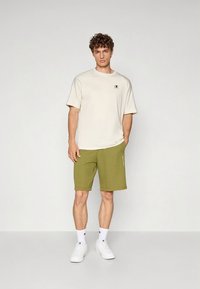 Ljusbeige t-shirt med kort ärm och logotyp, kombinerad med olivgröna shorts. Vita sneakers kompletterar den avslappnade outfiten.
