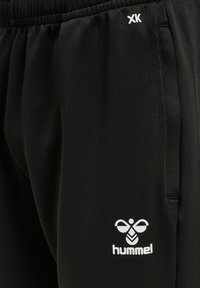 Svarta träningsbyxor gjorda av ett slätt material, med en vit "hummel"-logotyp på vänster lår och ett elastiskt midjeband med en subtil textur.