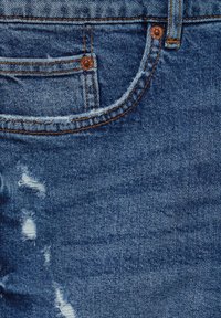 Tessuto denim blu con aree sfilacciate e un leggero invecchiamento. Presenta rivetti in rame e cuciture arancioni lungo le cuciture e le tasche.
