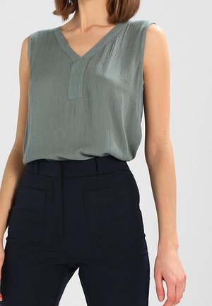 Vrouw draagt een mouwloze saliegroene V-halsblouse in een zwarte high-waisted broek, staand tegen een effen lichte achtergrond.