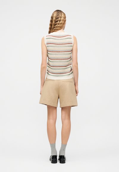 Γυναίκα με κυματιστά ξανθά μαλλιά που φοράει μπεζ σορτς, γκρίζες κάλτσες και μαύρα παπούτσια, ντυμένη με αμάνικη ριγέ πλεκτή μπλούζα, όρθια με την πλάτη γυρισμένη.