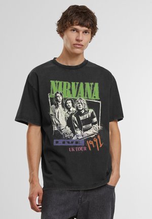 Jeune homme portant un t-shirt noir du groupe Nirvana avec un motif de la tournée britannique de 1991 et un jean noir sur fond uni.