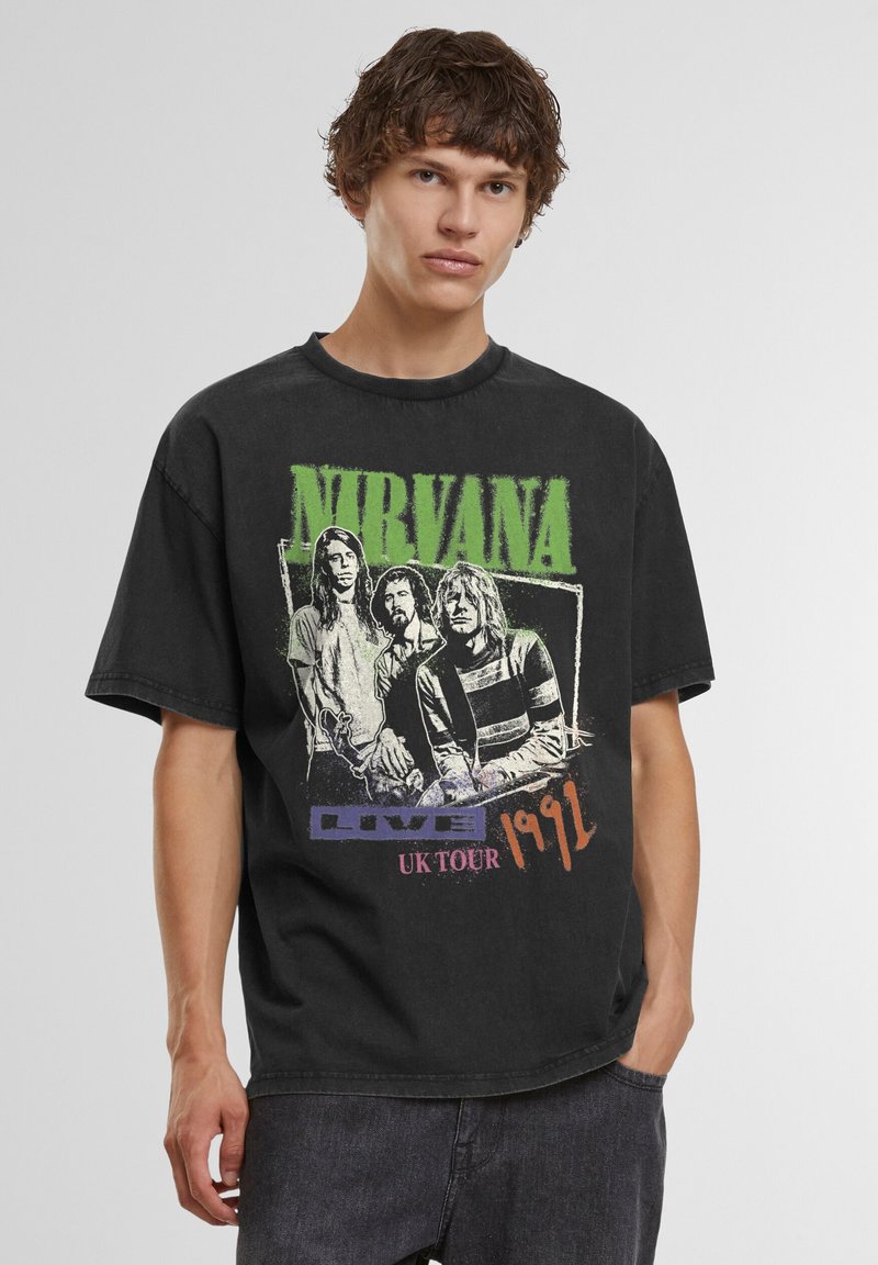 Jeune homme portant un t-shirt noir du groupe Nirvana avec un motif de la tournée britannique de 1991 et un jean noir sur fond uni.
