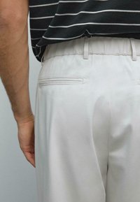 Uomo che indossa pantaloni elastici grigio chiaro e una camicia nera con righe bianche orizzontali, mostrato dalla vita in giú.