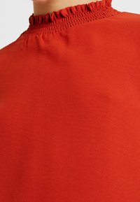 Haut rouge avec un col haut en texture froncée et un tissu lisse et léger. La vue en gros plan met en avant le matériau et la couleur.