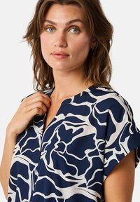Marinebluse mit weißem abstraktem Blumenmuster, kurzen Ärmeln und einem V-Ausschnitt. Glatter Stoff, eng anliegendes Design an den Schultern.