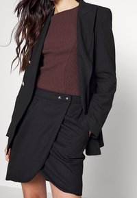 Svart blazer med guldknappdetaljer, kombinerad med en ribbad burgundytopp och en svart inslagskjol med silverdetaljer på midjebandet.