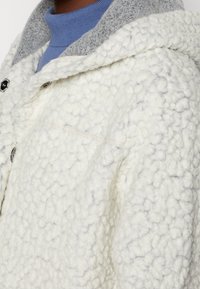 Chaqueta de forro polar blanco con un acabado texturizado y esponjoso, que cuenta con un forro de capucha gris, botones a presión y un sutil detalle de costura en el pecho.