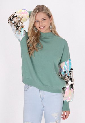 Grüner Pullover mit hohem Kragen und puffigen Ärmeln, geschmückt mit mehrfarbigen Pailletten. Hellblaue, ausgebleichte Jeans runden das Outfit ab.