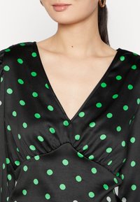 Abito di raso nero con pois verdi, scollo a V e dettagli arricciati in vita, con maniche lunghe. Texture liscia.