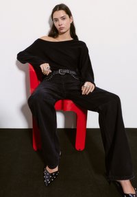 Črn oversized pulover, raztrgane črne široke hlače z srebrnimi zakovami, črne pointed heels s zakovami, sedi na rdečem stolu.