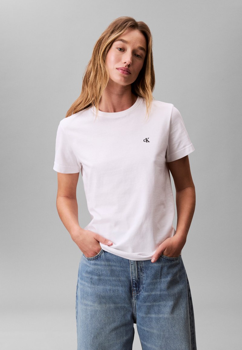 Weißes Baumwoll-T-Shirt mit Rundhalsausschnitt und kurzen Ärmeln, mit einem kleinen schwarzen "cK"-Logo auf der linken Brust. Kombiniert mit hellblauen Denim-Jeans.