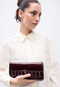 Žena v priesvitnej bielej blúzke s kvetinovým vzorom drží tmavý bordový kožený listový kabelku so striebornými očkami a detailom pracky.