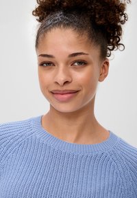 Jasnoniebieski sweter z dzianiny z okrągłym dekoltem, teksturowanym materiałem i prążkowanym wykończeniem. Modelka ma kręcone włosy i patrzy w przód.
