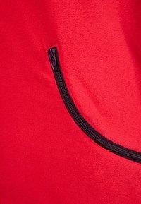 Červený fleecový materiál s křivkovou kapsou a černým zipem. Textura je měkká a detail se zipem přidává funkčnost.