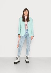 Blazer verde menta, suéter de gola alta rosa com padrão de corações, jeans de cintura alta azul claro, botas brancas com atacadores e uma pequena mala rosa.