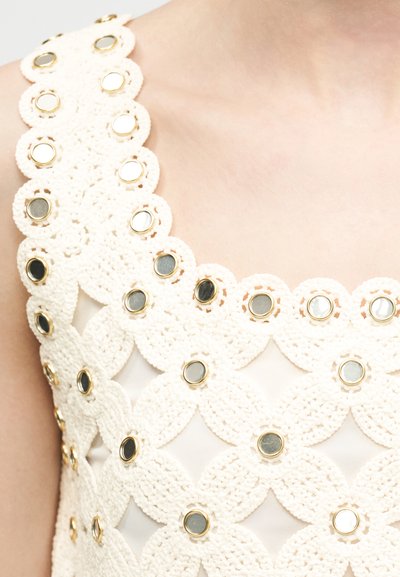 Vêtement blanc en crochet avec des ornements circulaires à bord doré et miroir, porté sur l'épaule et le haut de la poitrine d'une personne.