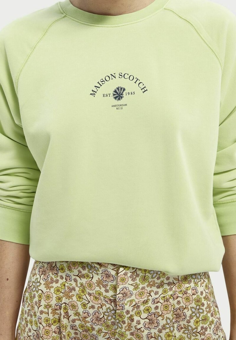 Persoon draagt een lichtgroene Maison Scotch trui met logo en een broek met bloemenpatroon in beige, groen en bruine tinten.