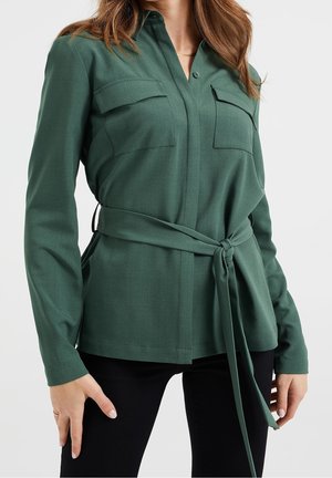 Femme portant une chemise longue à manches longues vert foncé ceinturée avec deux poches poitrine, associée à un pantalon noir, sur fond blanc.