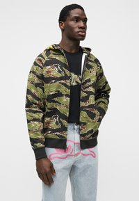 Ζακέτα με φερμουάρ σε αποχρώσεις του πράσινου, μαύρου και καφέ με ριμπ μανσέτες. Φορεμένη πάνω από μαύρο μπλουζάκι και συνδυασμένη με τζιν ανοιχτού χρώματος με ροζ λεπτομέρειες.