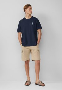 Marineblauwe t-shirt met korte mouwen en palmboomafbeelding op de borst, gecombineerd met lichtbeige cargoshorts enbruine slidesandalen.