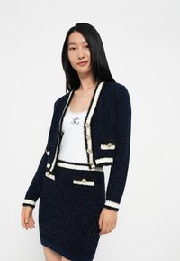 Cardigan lavorato a maglia navy con dettagli crema, bottoni dorati e tessuto texturizzato. Abbinato a un top bianco con logo. Design aderente.