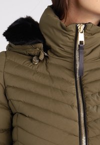 Olivově zelená prošívaná bunda s vysokým límcem, zlatým zipem, černou kapucí s kožušinovým lemem a kovovým zapínáním v oblasti ramene.