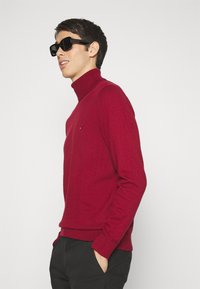 Raudonas turtleneck megztinis su raukšlėta apdaila, dėvimas su juodais kvadratiniais akiniais nuo saulės. Modelis stovi šonu, rankomis kišęs į kišenes, prieš paprastą foną.