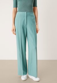 Breda byxor i ljus teal-tyg, med en framsida som har veck och en ren klippning med en mjuk finish. Parat med vita sneakers.