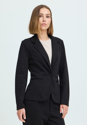 Noor naine, kellel on sirged õlgadeni ulatuvad juuksed, kannab musta bleiserit valge topi kohal ning seisab ühevärvilise heleda tausta ees.