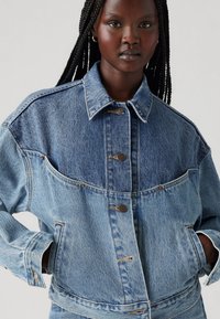 Jeans jakna s dvobarvnim modrim dizajnom, klasičnim ovratnikom, zapiranjem na gumbe in stranskimi žepi. Lesena tekstura dodaja vizualno privlačnost.