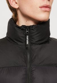 Schwarze gepolsterte Jacke mit hohem Kragen, ausgestattet mit einem Frontreißverschluss und einem Logodetail am Reißverschluss. Glatte Textur und minimalisches Design.