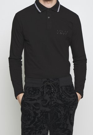 Homme portant un polo noir à manches longues avec des rayures blanches sur le col et un pantalon noir à motifs avec cordon, mains dans les poches.