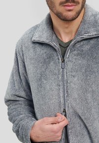 Chaqueta de fleece gris con cremallera y un patrón texturizado y acanalado. Cuenta con cremallera frontal y mangas largas.