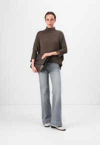 Poncho en tricot marron avec col roulé, coupe décontractée, associé à des jeans évasés bleu clair. Tient une pochette marron foncé avec un fermoir.
