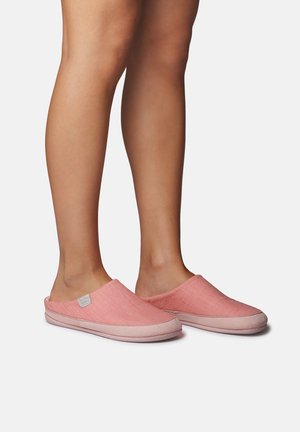 Jalad ja alakehad, mis kannavad roosaid klanitud sussisid, mille pealmine osa on pehme kangaga ja heleroosa tald, ühtlase tausta vastu.