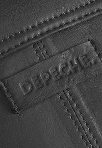 Černý kožený produkt s texturovaným povrchem, prošitými okraji a výraznou embosovanou nálepkou s nápisem "DEPECHE" v kapitalizovaných písmenech.