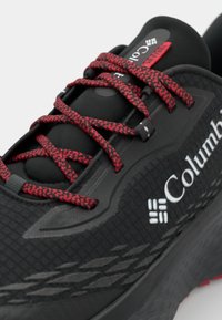 Shoe sport negru cu o suprafață texturată, șireturi cu model roșu și negru și logo alb. Are un guler căptușit și un sistem de șireturi sigur.