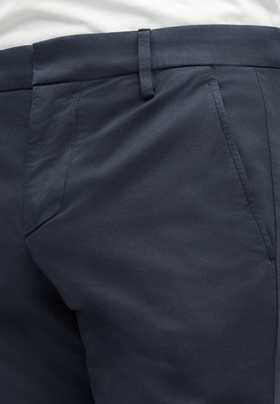 Εγγύς πλάνο σκούρων ναυτικών παντελονιών με ορατή μέση, θηλιά ζώνης και εμπρόσθιες τσέπες, φορεμένα με λευκό πουκάμισο που έχει μπει μέσα.