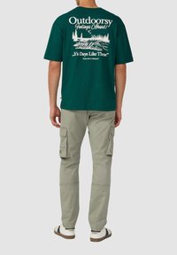 Groene katoenen T-shirt met een witte bedrukte buitenomgeving en tekst op de achterkant, gecombineerd met grijze cargobroeken en witte sneakers.