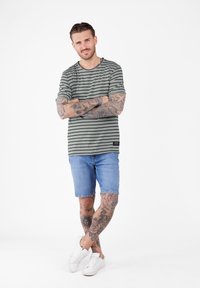 Hombre con tatuajes que lleva una camiseta de rayas, pantalones cortos de mezclilla y zapatillas blancas, de pie con los brazos y las piernas cruzadas sobre un fondo blanco.